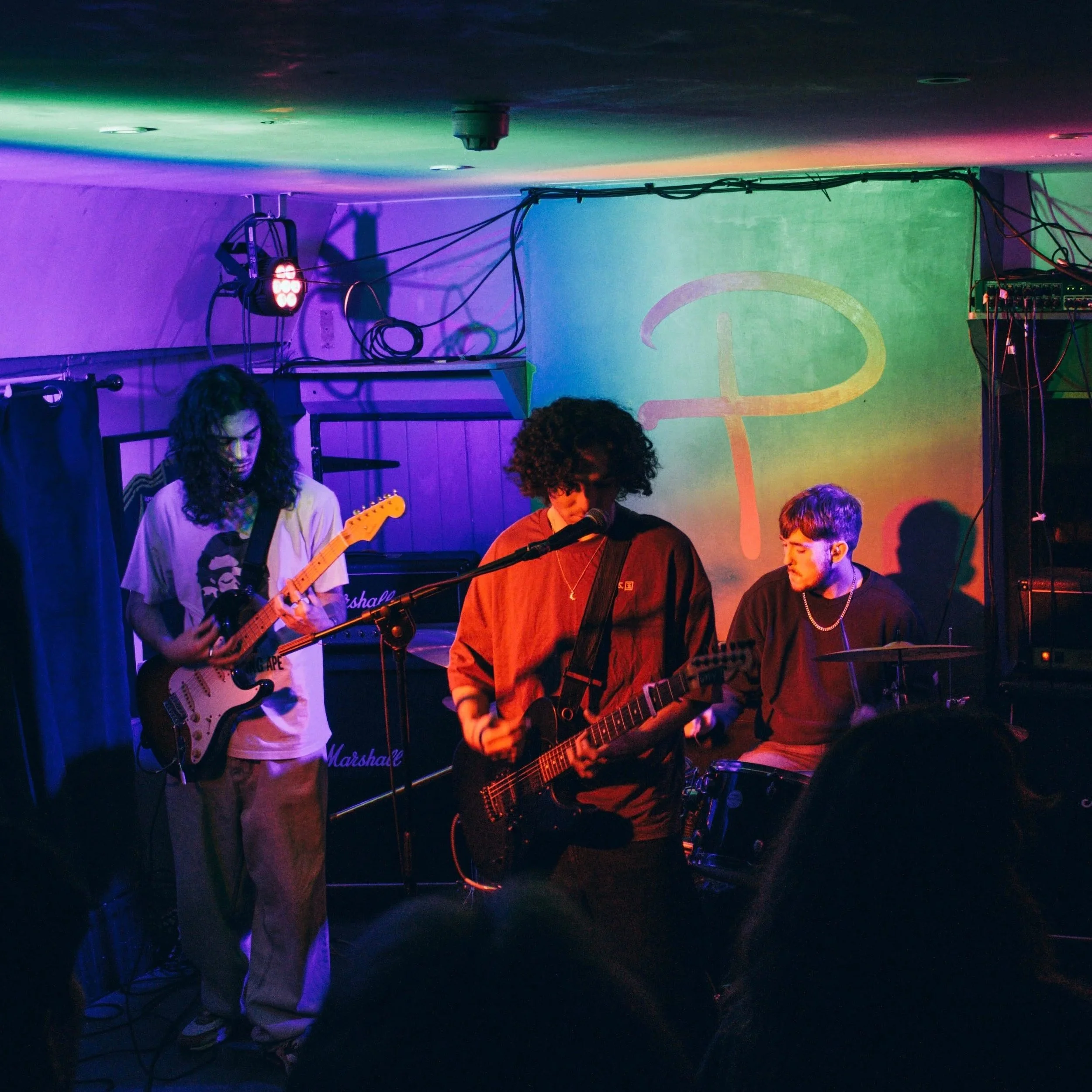 Three musicians performing on a stage with colorful lighting, one playing a guitar, another with an electric guitar, and the third on drums, with a gradient backdrop and sound equipment visible.