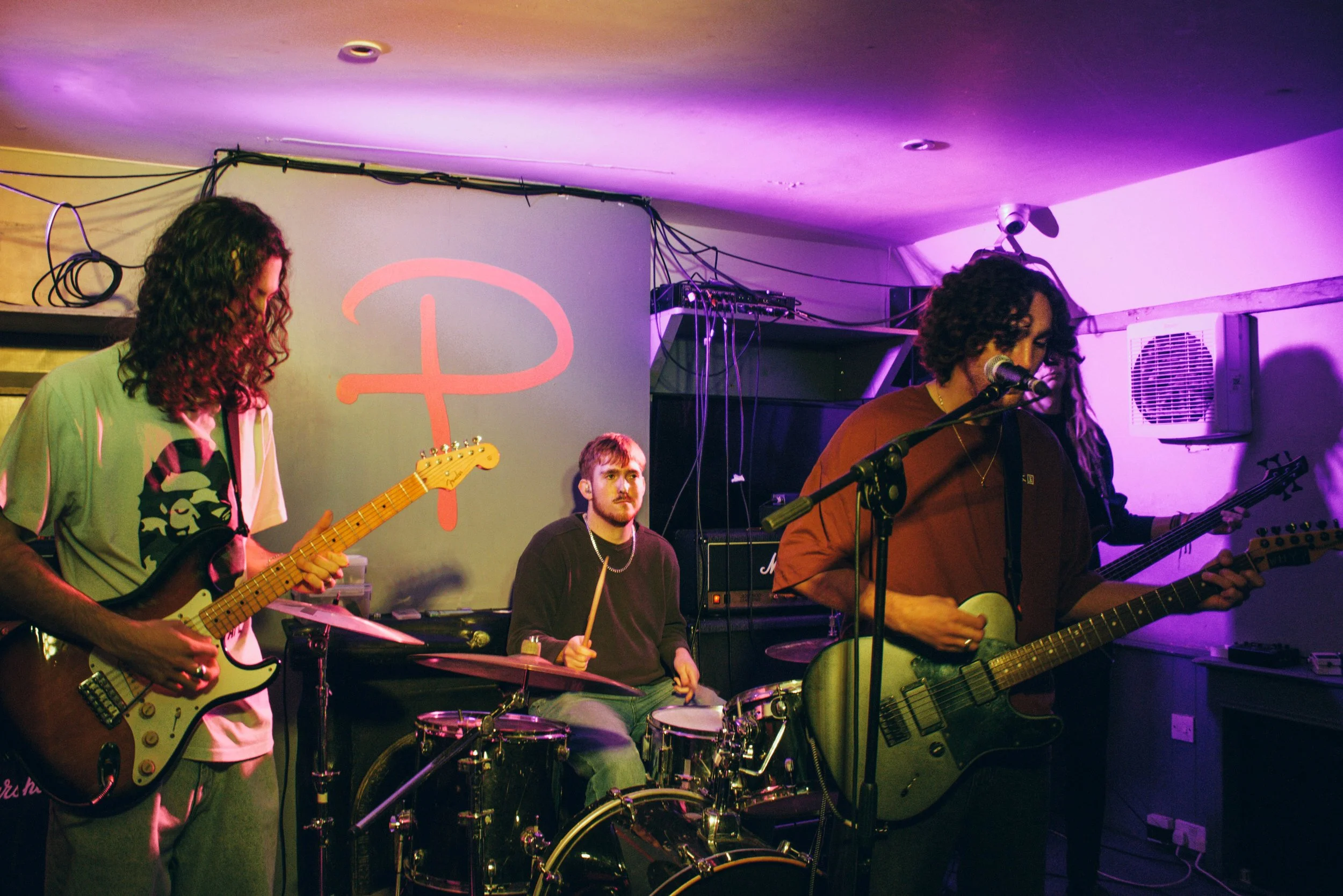 Four musicians performing on stage, with two guitarists and a drummer, in a room with purple lighting and a "D" logo on the wall.