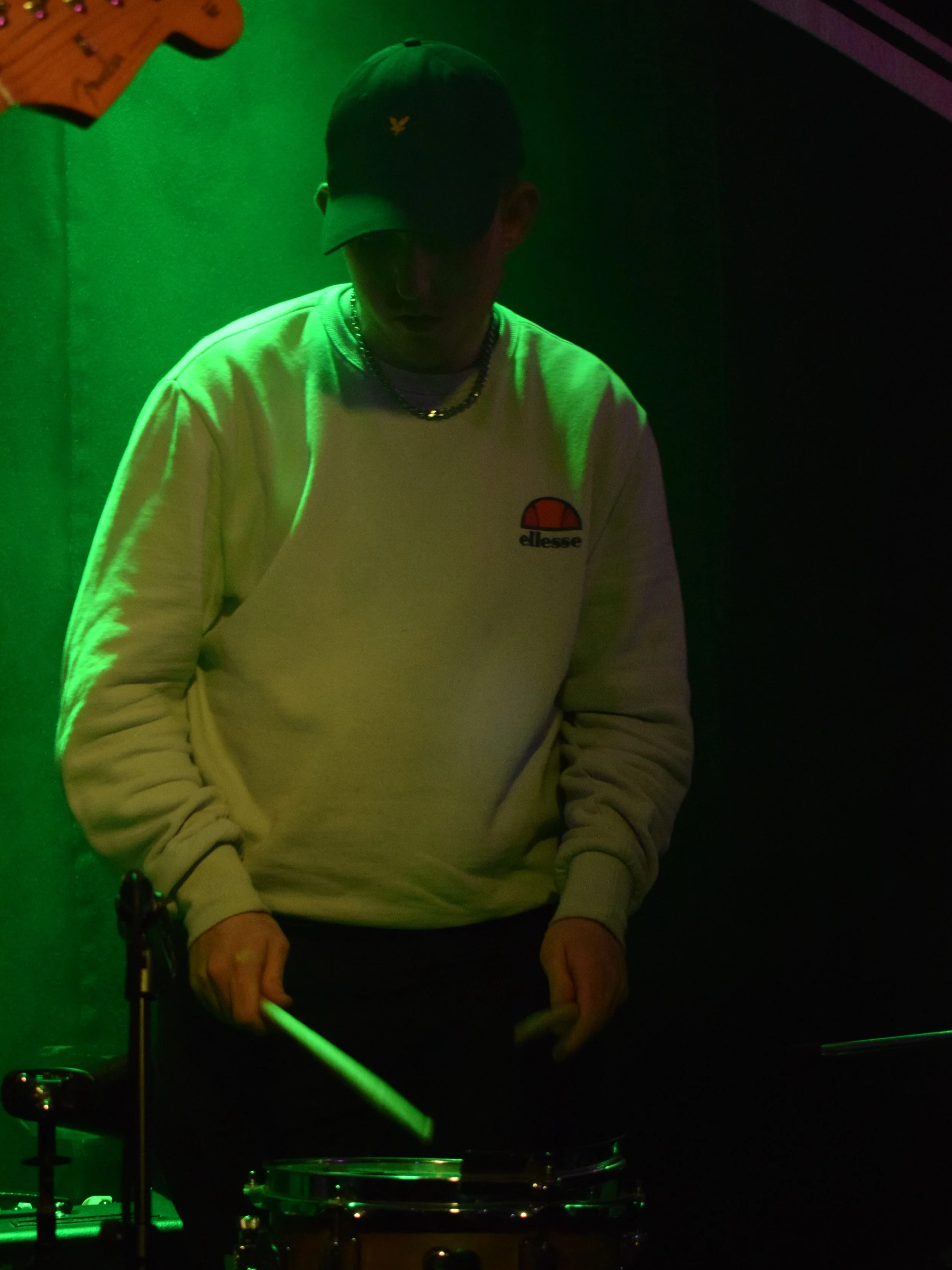 Young person wearing a dark cap and a white sweatshirt with an orange and black logo, standing in front of a green-lit background, holding drumsticks near a drum.