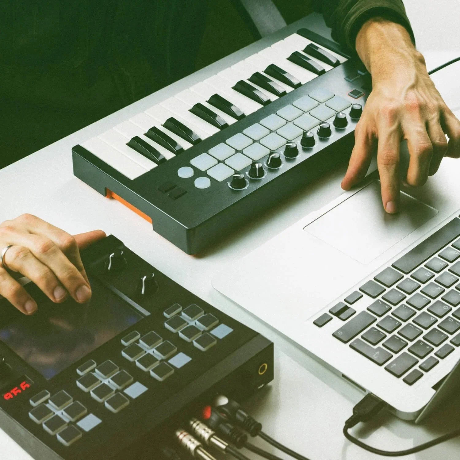 Hands working with music production equipment, including a small MIDI keyboard, a laptop, and a pad controller on a white table.