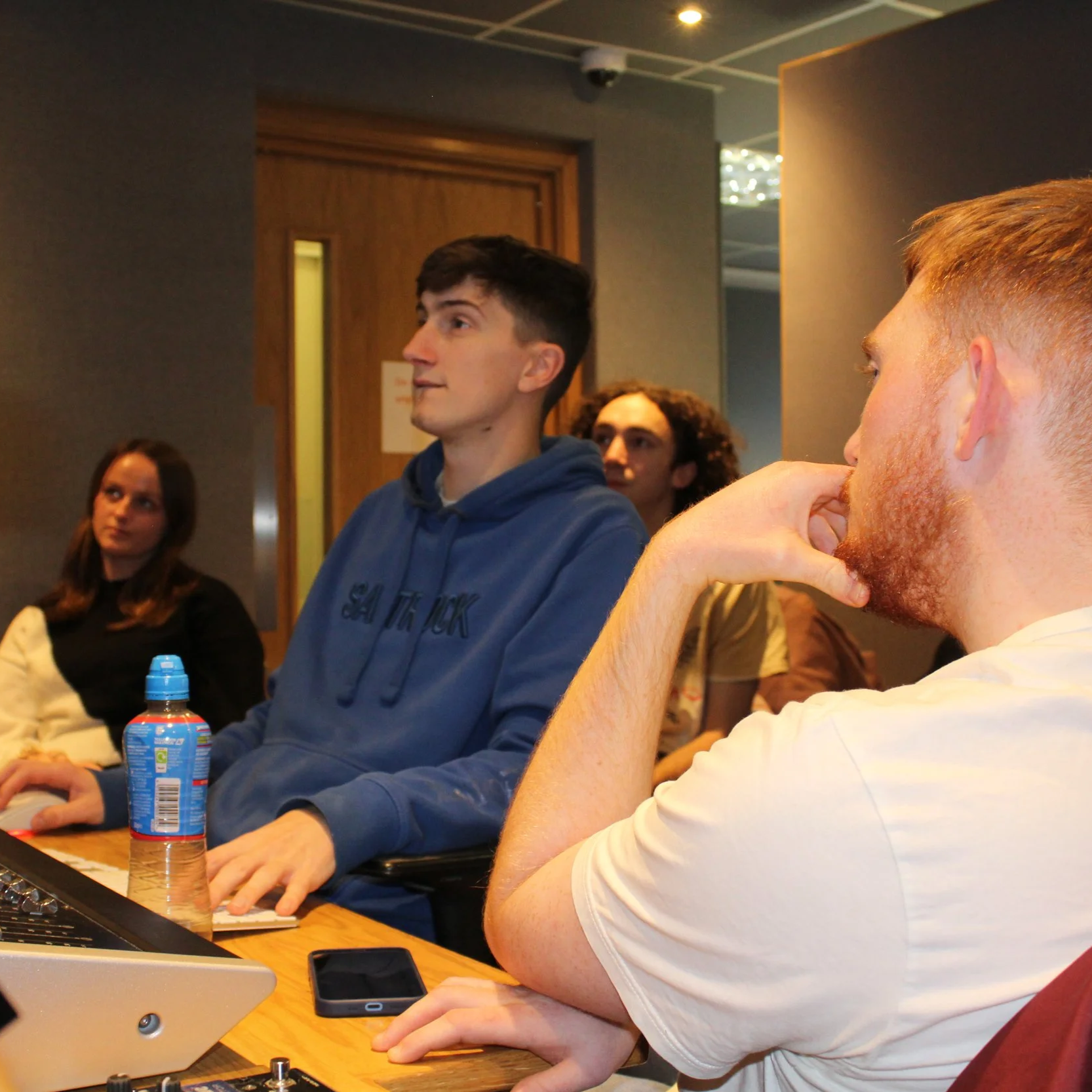 A group of young adults within a music studio, recording vocals for "Comedown", Sleepflower's debut single. They are sitting around a mixing desk with a laptop, a water bottle and a phone. Lewis Cole. Harry Amos. Molly Smith. Henry Hockings.