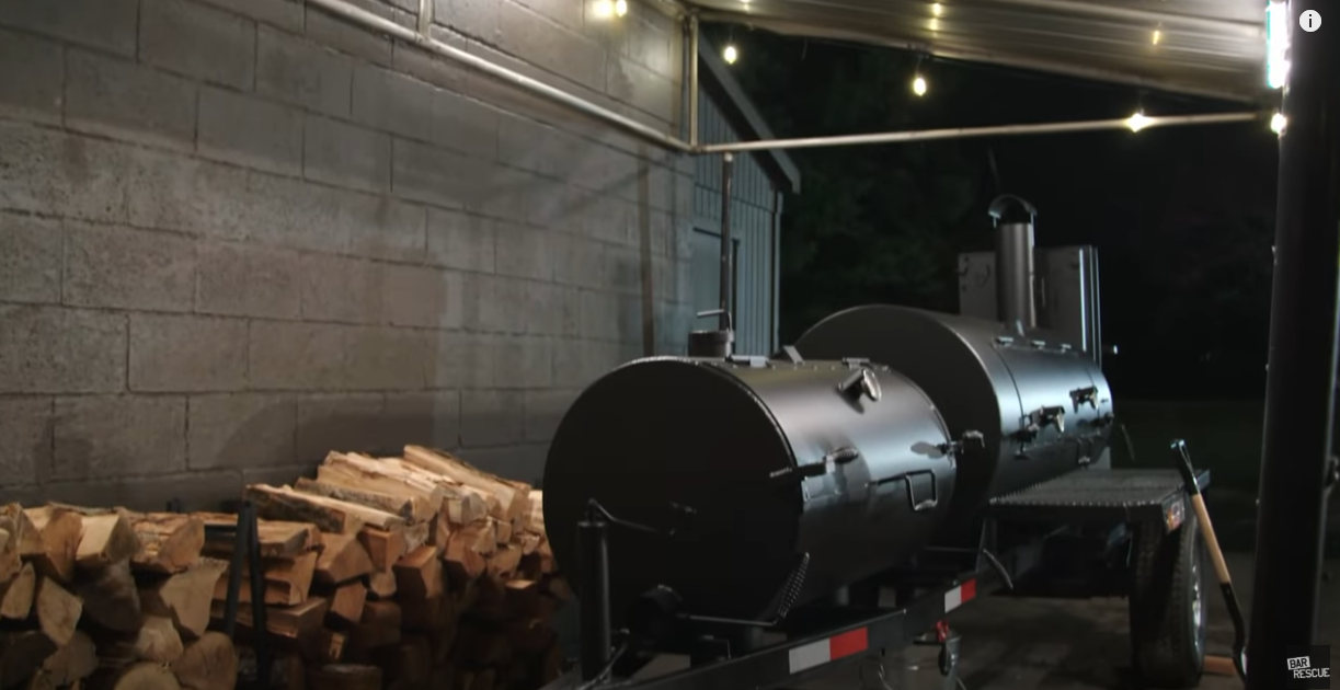 A black outdoor smoker on a trailer parked near a brick wall, with a pile of chopped firewood nearby, under a covered area at night.