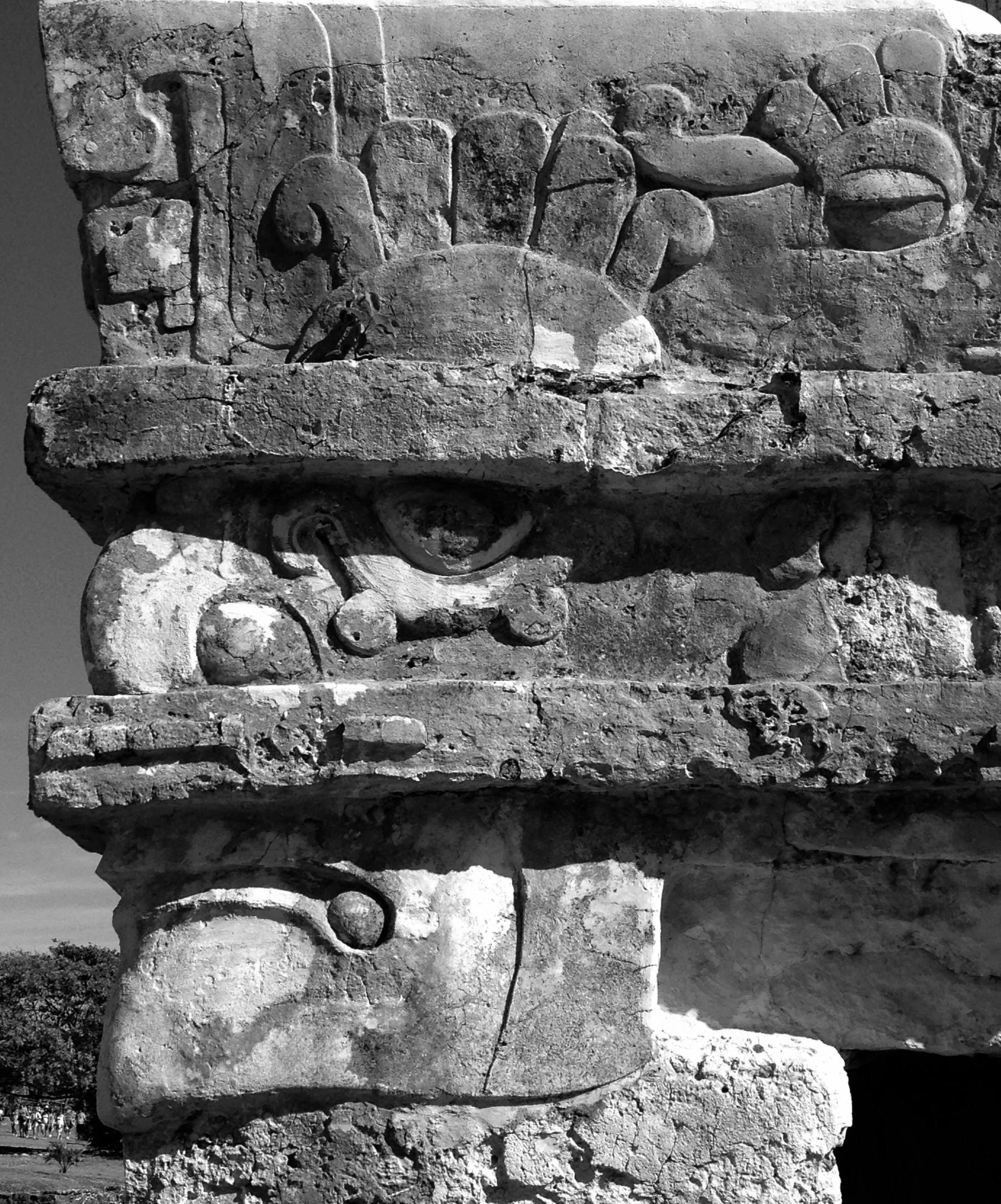 Close-up of ancient carved stone ruins with multiple layers of weathered stone blocks, some with inscriptions and circular carvings, set against a clear sky with a distant landscape.