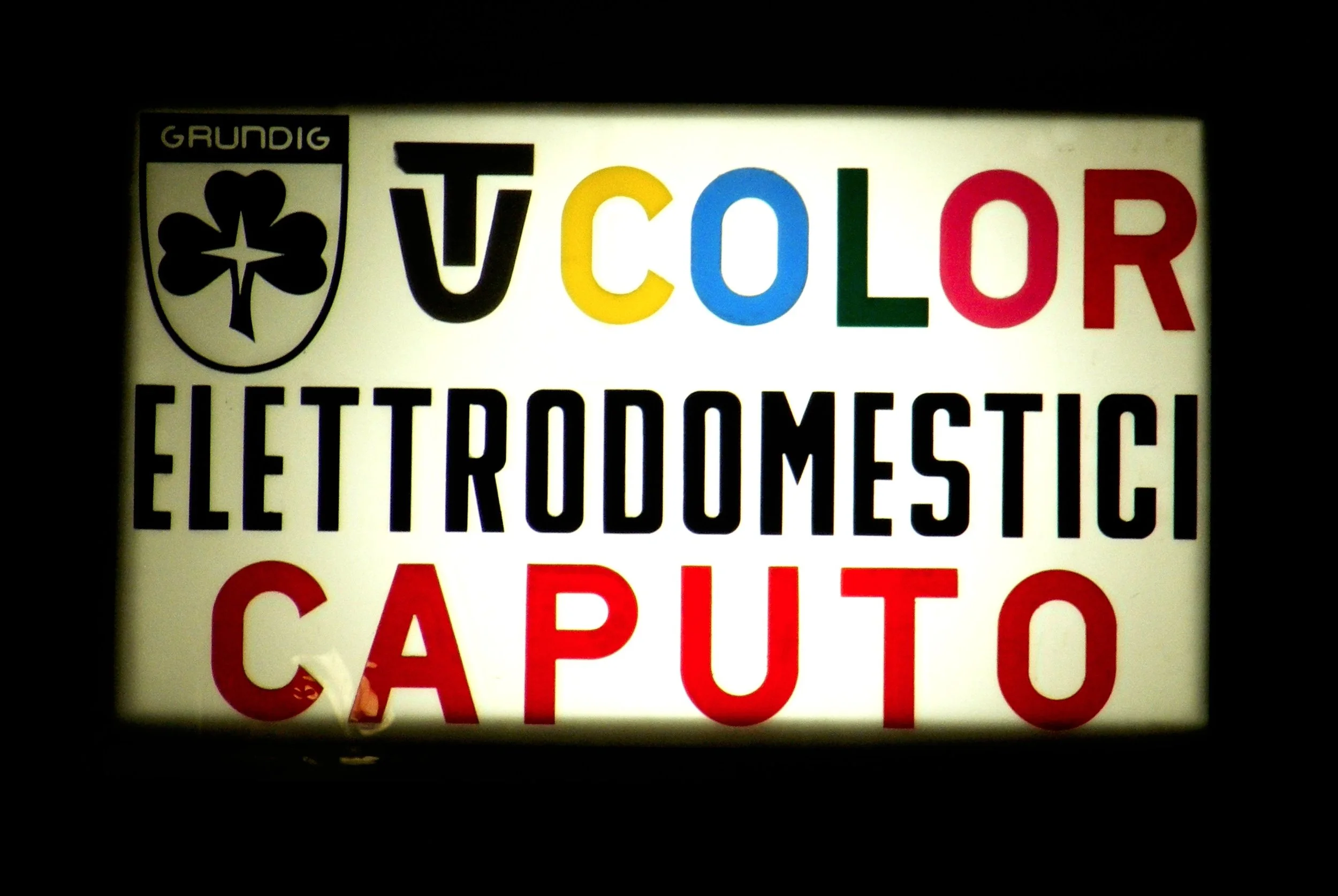 Illuminated sign displaying the logo of Grundig, with a black shield and a three-leaf clover, and the words 'TV COLOR' in multicolored letters and 'ELECTRODOMESTIC' in black text below.