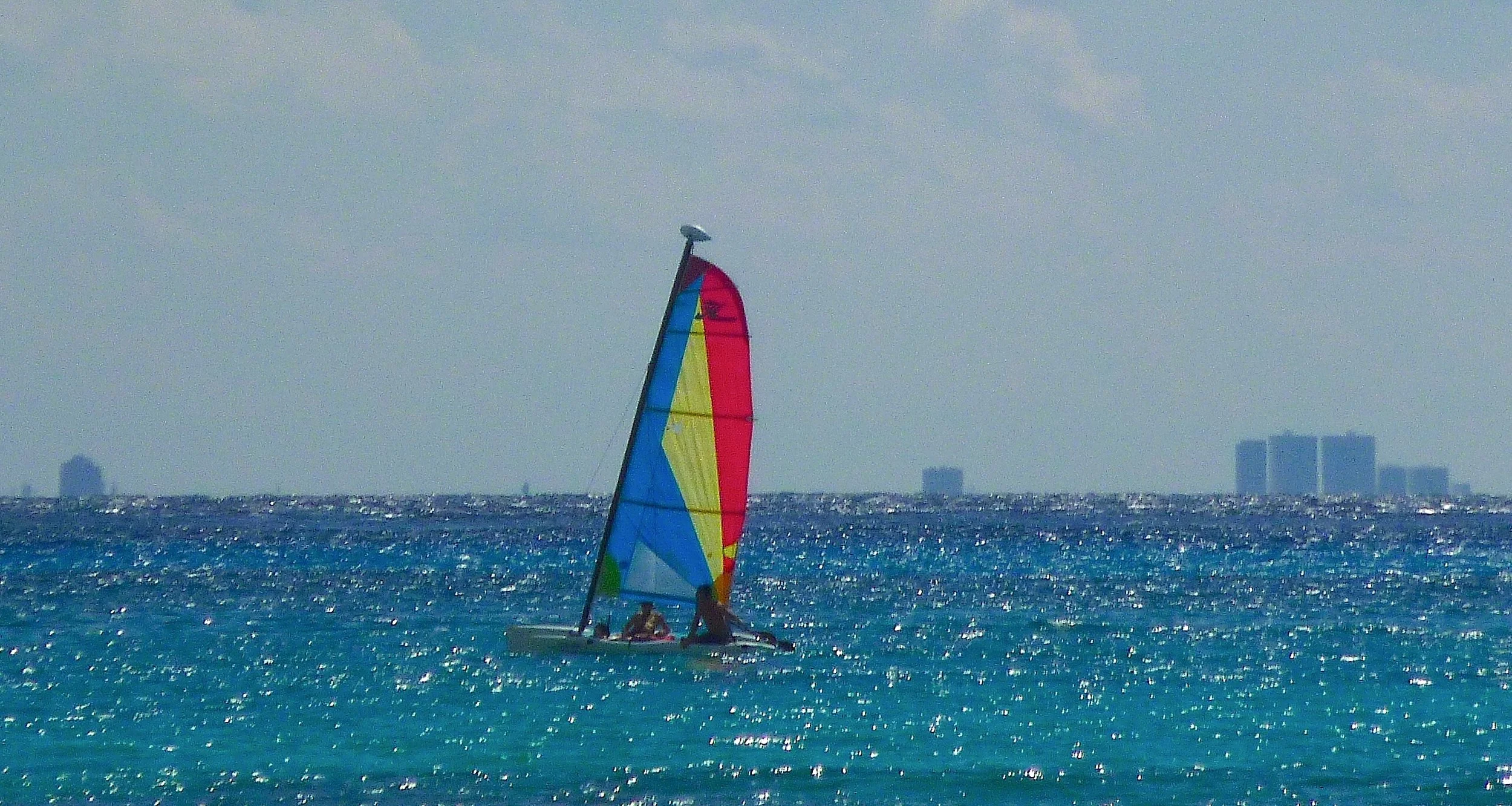 A colorful sailboat with a rainbow-colored sail riding on the ocean with a city skyline in the distance under a cloudy sky.
