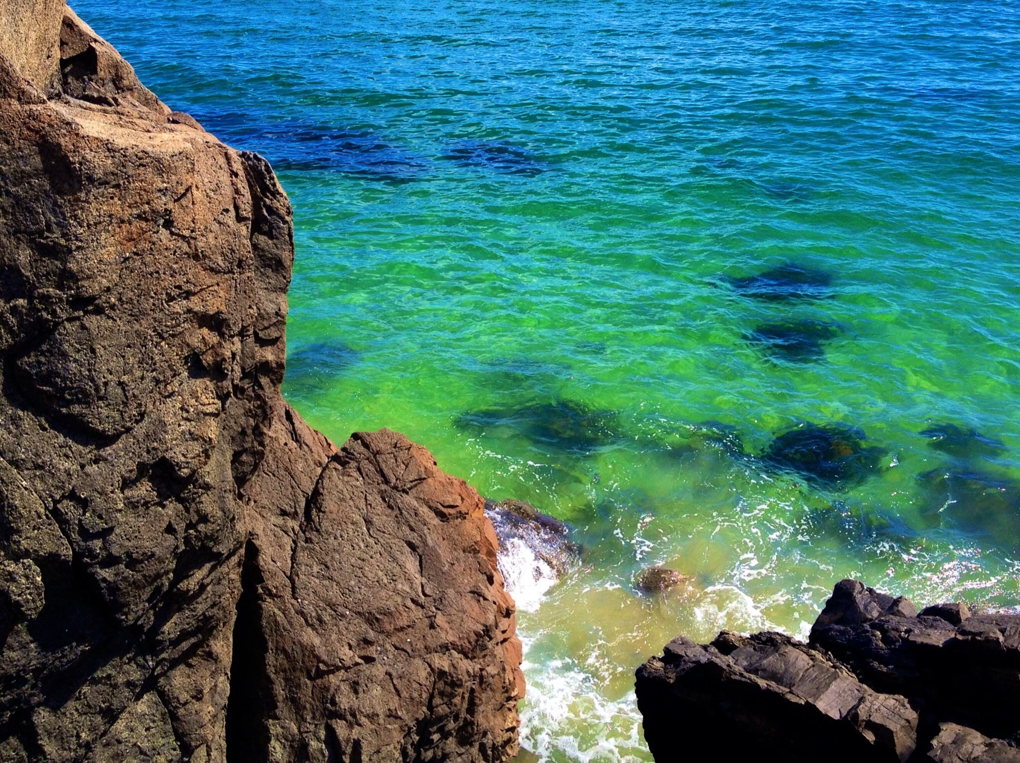 Large dark brown rocks on a rocky shoreline with clear green and blue ocean water.