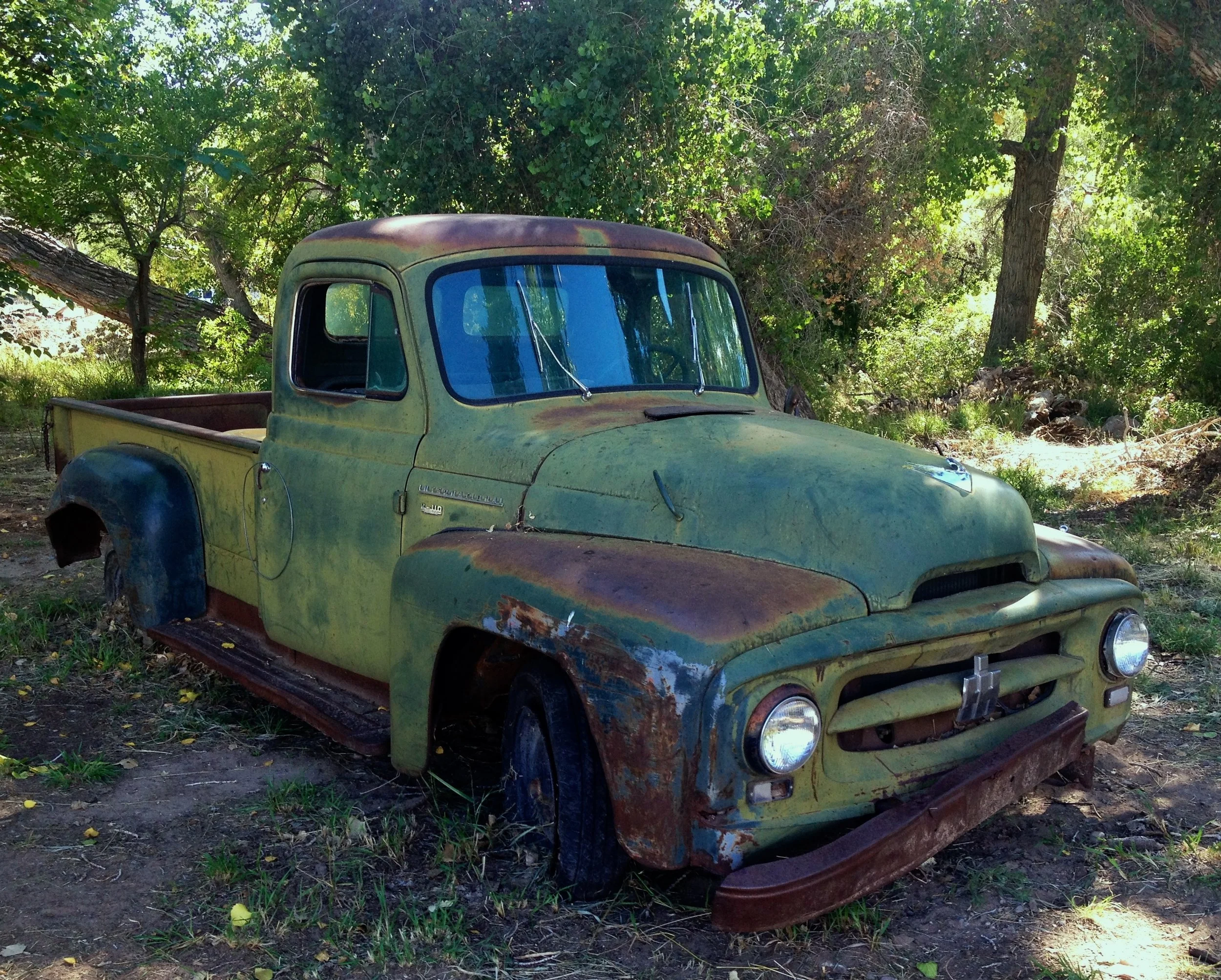 An old, rusty green pickup truck parked in a wooded area surrounded by trees and grass.