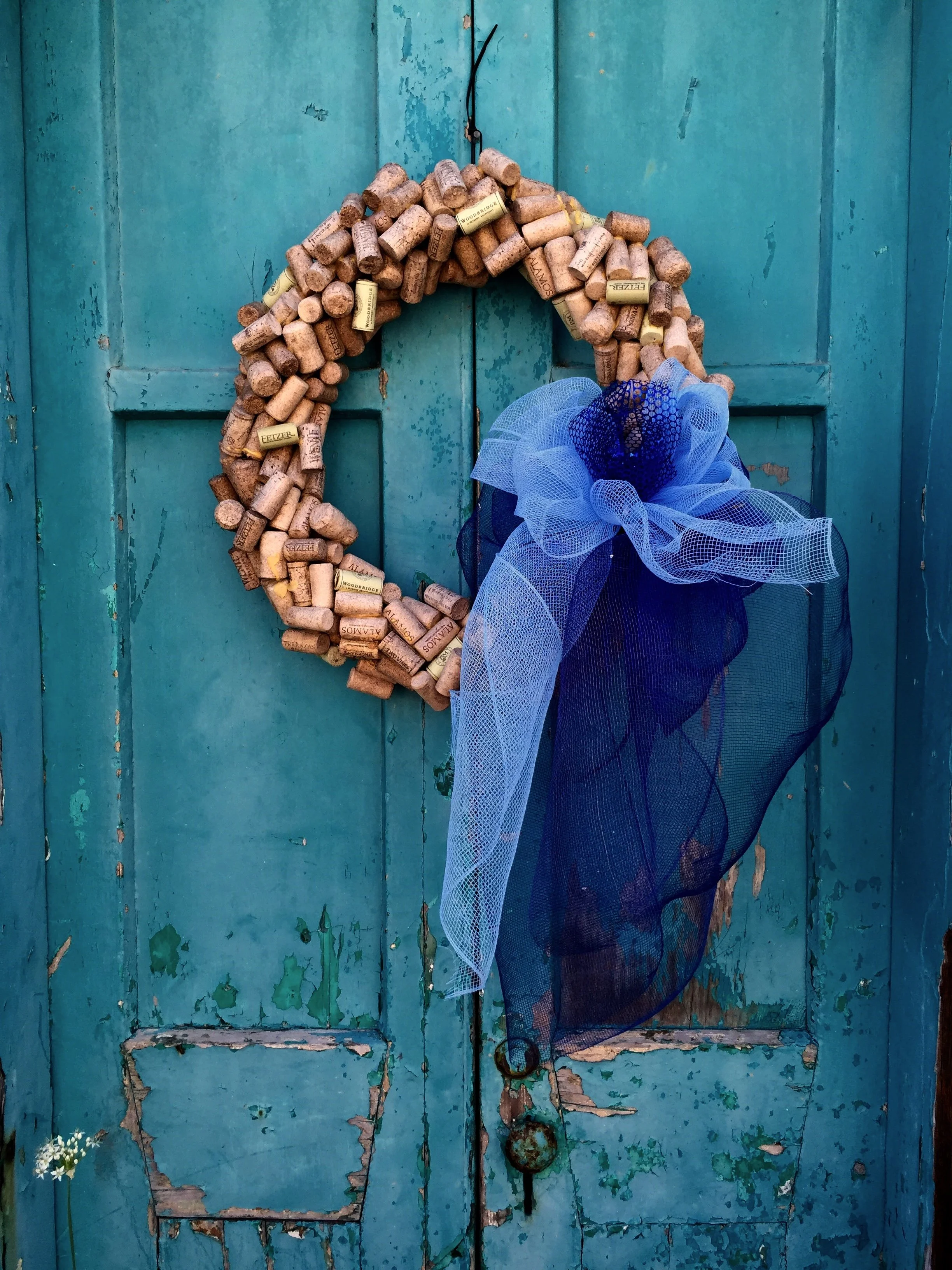 A blue wooden door with peeling paint, decorated with a heart-shaped cork wreath and a large sheer blue bow, with a small white flower bouquet on the left side.