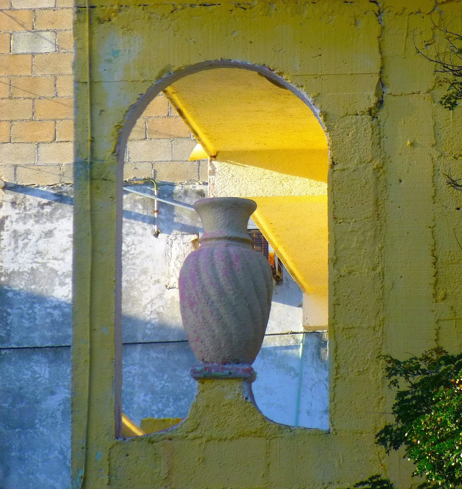 A yellow brick wall featuring an arched opening with a decorative vase or urn placed on a ledge. The area outside the arch shows a textured white wall and some vegetation on the lower right corner.
