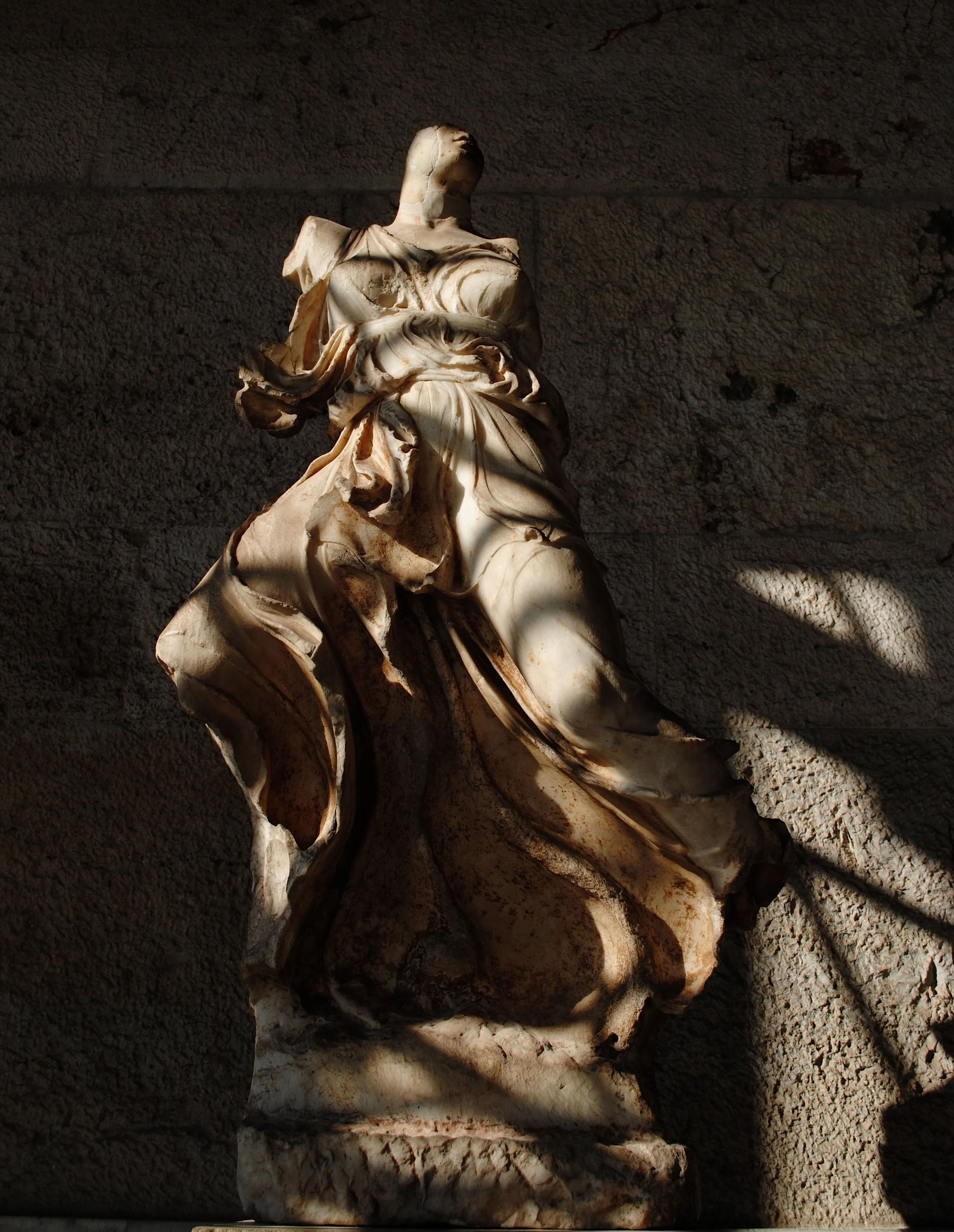 Ancient marble statue of a seated figure against a textured stone wall, with dramatic lighting highlighting details of the statue's drapery and form.