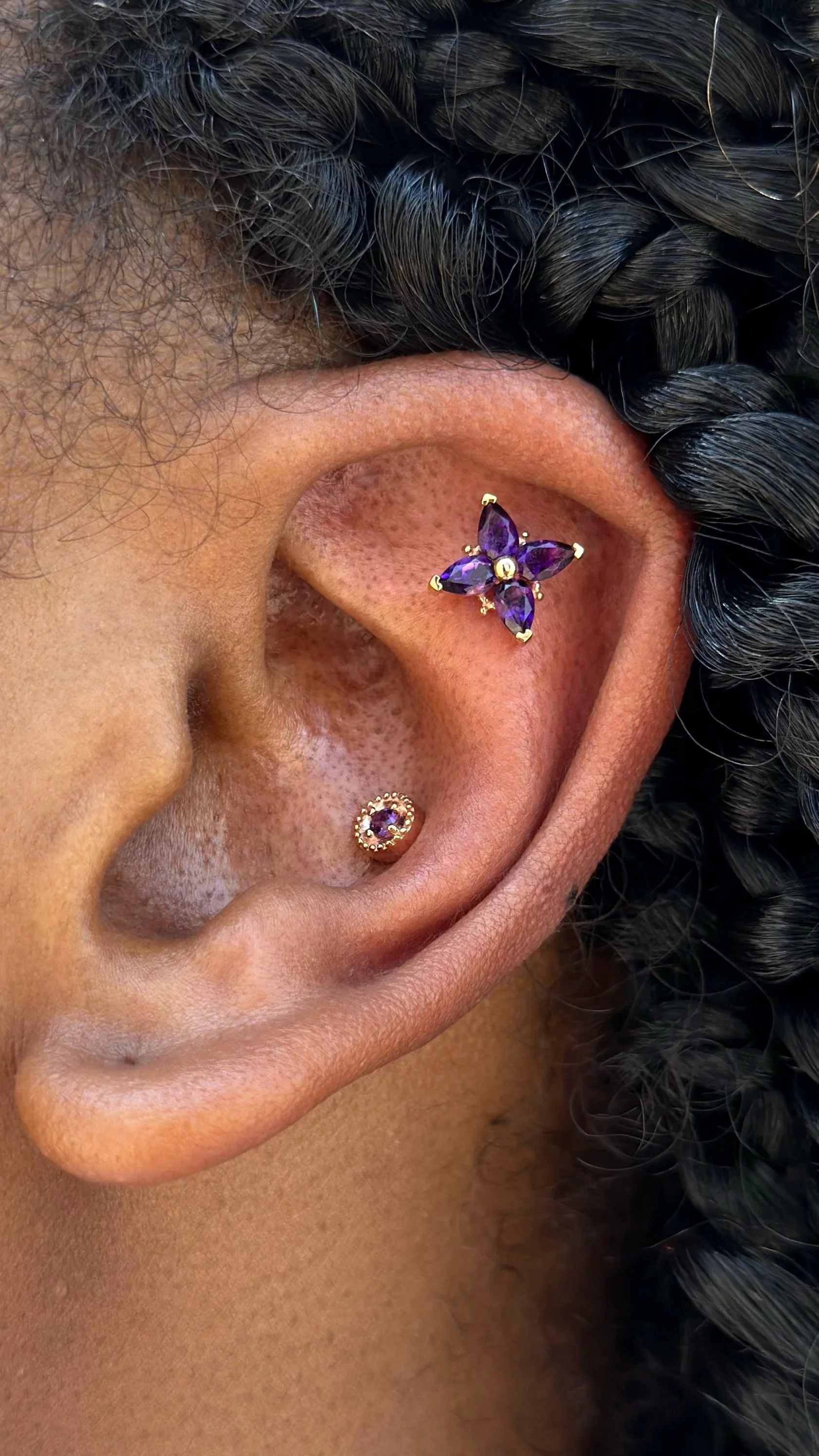 Close-up image of a person's ear with two purple Amethyst gemstone earrings, one in a flower shape and the other in a circular setting, both set in solid gold.