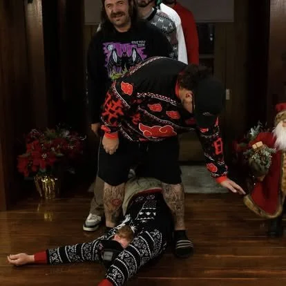 Two men, one standing and one kneeling. The man standing is smiling and wearing a black cap, black shorts, and a colorful shirt. The kneeling man is on the floor, wearing a black and white patterned sweater, and has a woman lying on his back. The background shows holiday decorations and a wooden floor.