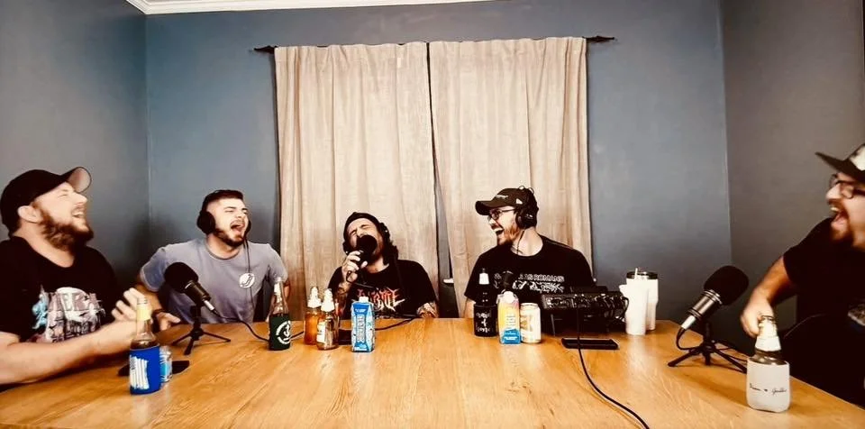 Five men sitting around a wooden table with drinks and microphones, laughing and talking in a casual setting, possibly recording a podcast.