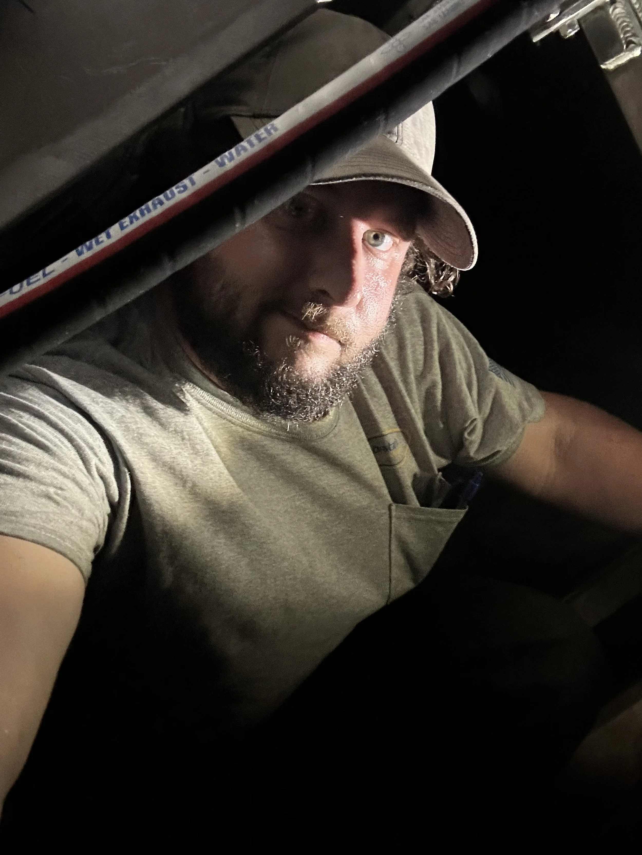 A man with a beard and mustache wearing a tan baseball cap and a gray t-shirt, looking into the camera from inside a dark space, illuminated by a light source.