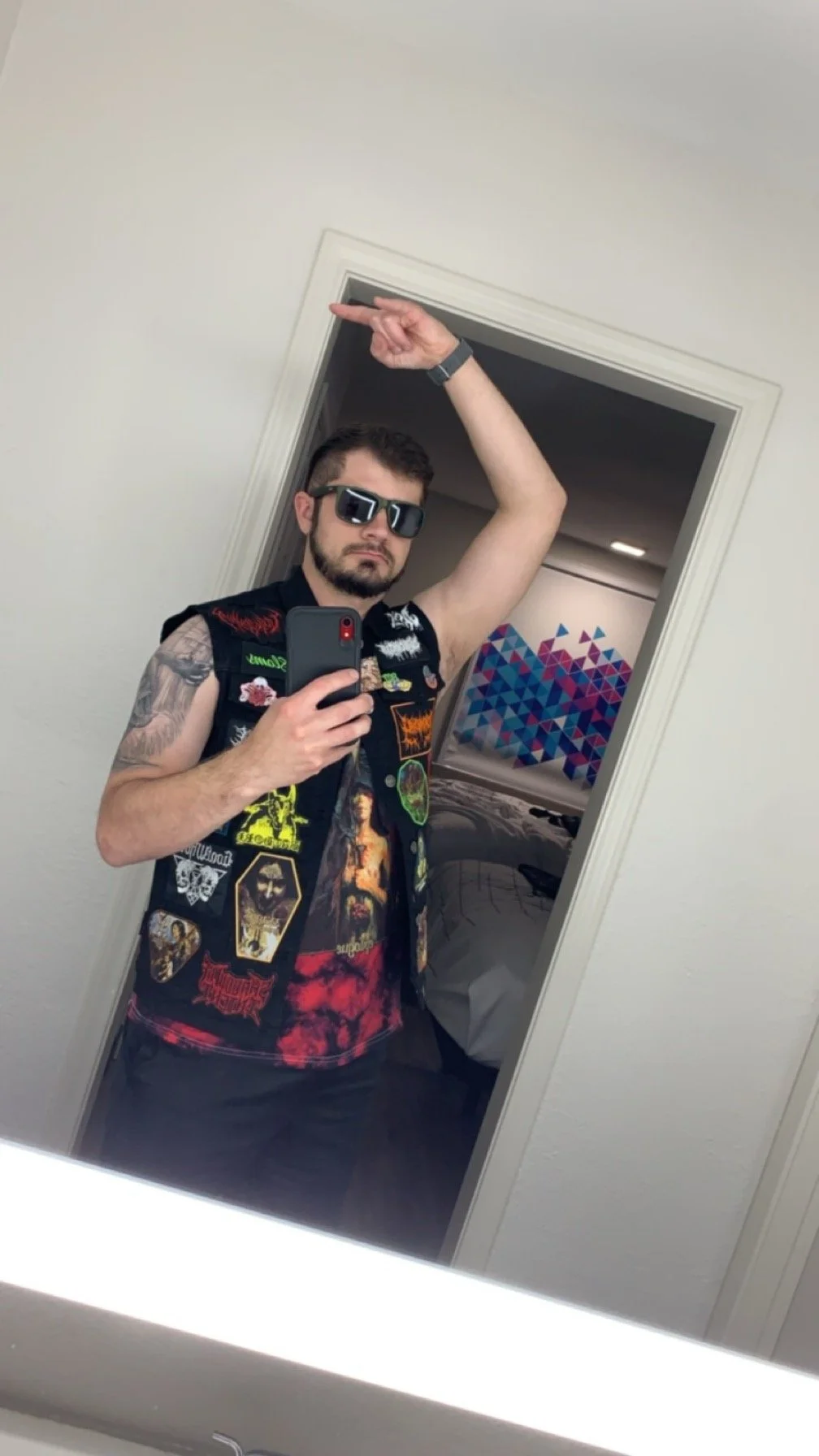 A man taking a selfie in a mirror, wearing sunglasses, a sleeveless vest with patches, and black pants. He has tattoos on his left arm and is making a pointing gesture with his right hand. The background shows a hotel room with a bed and a decorative wall art.