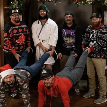 Five men standing indoors, with two men doing push-ups in front. Some are dressed in Christmas sweaters, and one is wearing a Santa hat. The setting appears festive.