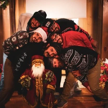 A group of seven people dressed in festive clothing, posing together with a Santa Claus figure in front of Christmas decorations.