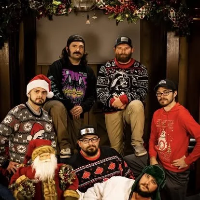 Five men posing in front of Christmas decorations, wearing holiday sweaters, with one in a Santa hat and another with a Christmas-themed beard. Santa Claus figure is seated in front.