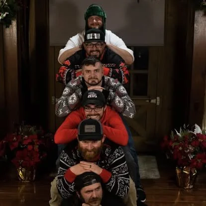 Six men sitting and posing in a vertical line, stacked one on top of the other, with holiday sweaters and festive decorations around.