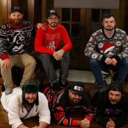 Six men posing indoors, wearing Christmas sweaters, some sitting on the floor and others kneeling behind them.