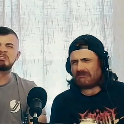 Two men sitting at a table with microphones, one wearing a baseball cap and headphones, the other not. Both are making expressive facial gestures.