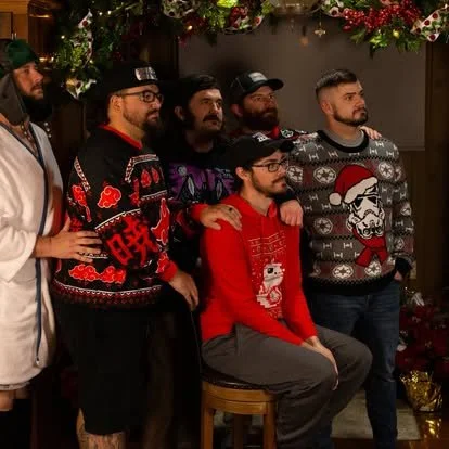 Five men posing for a festive photo in front of holiday decorations, wearing Christmas sweaters with holiday-themed designs.