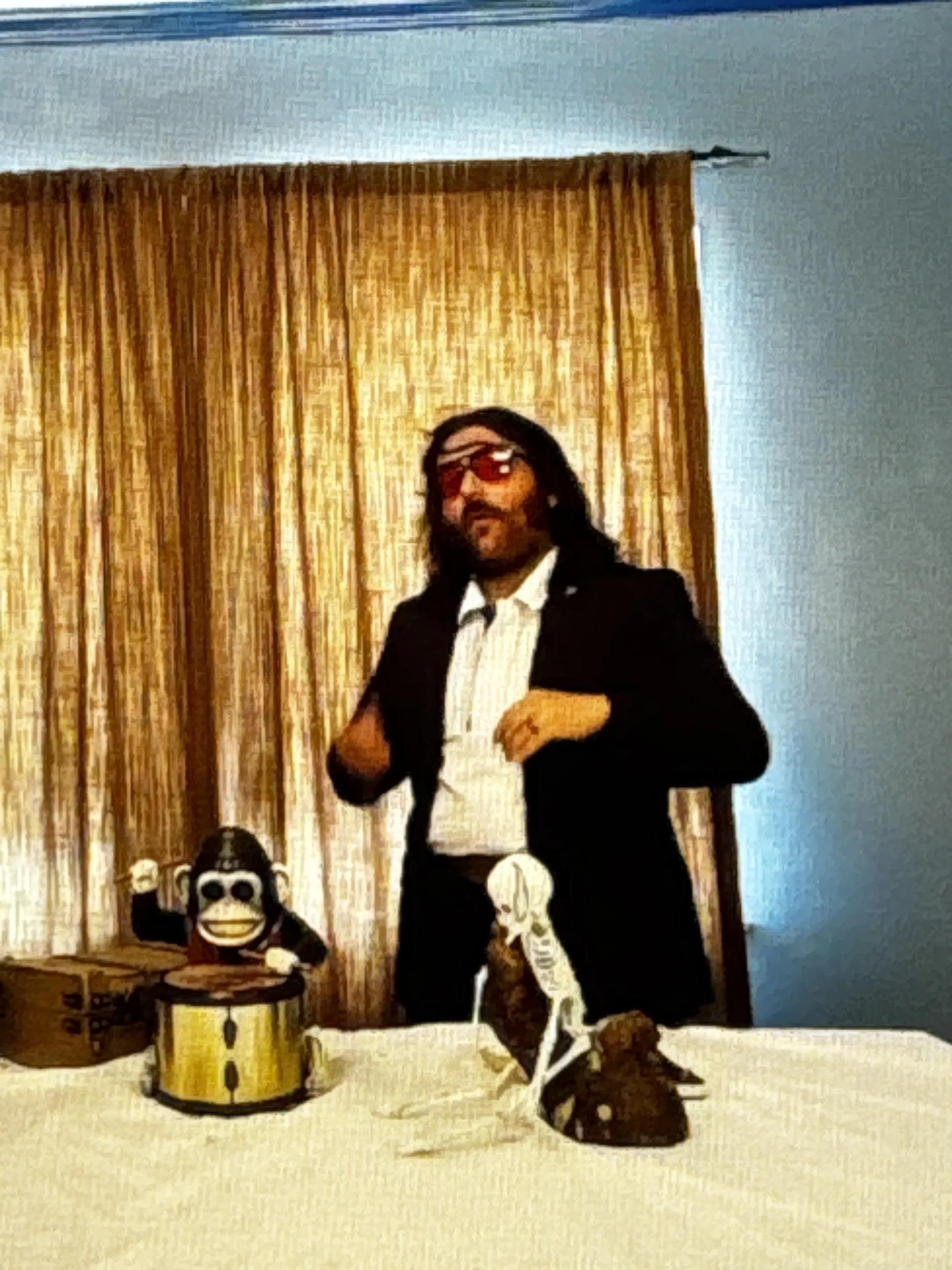 Person with long hair and sunglasses in a suit standing behind a table with monkey and ape figurines, a drum, and a skeleton model, in front of gold curtains.