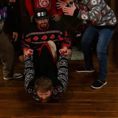 Group of people celebrating during a holiday gathering, with one person lying on the floor holding another person upside down, all wearing Christmas sweaters.