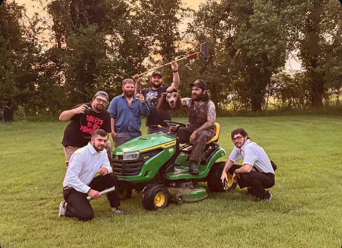 Group of six men on a grassy field with a green John Deere riding lawn mower, one of whom is wearing a horse mask and holding a spear. The men are posing for a photo during sunset, with trees in the background.