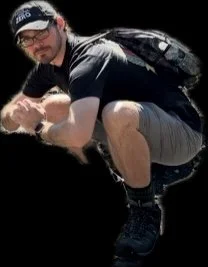 A man crouching down, wearing a black cap, glasses, a black T-shirt, gray shorts, and a backpack, with a black background.