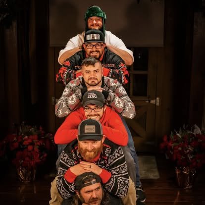 Five men in Christmas sweaters and hats forming a human pyramid indoors, with Christmas decorations and flowers in the background.