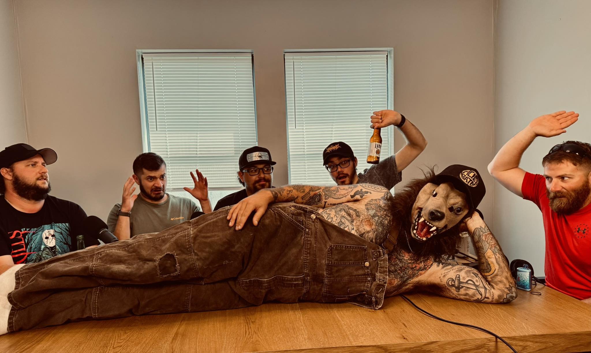 A group of six men around a table, with a person wearing a dog mask and tattoos lying on the table in front of them, holding a beer bottle and making celebratory gestures.