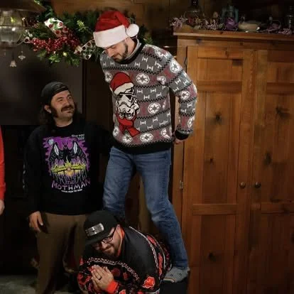 Three men celebrating Christmas indoors. One man in a Santa hat and Christmas sweater stands on a stool, another man in a Batman shirt and black cap stands nearby, and a third man in a red and black sweater is crouched down, holding a small gift.