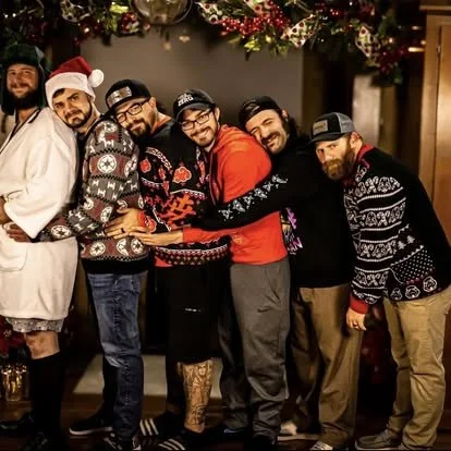 Group of six men standing close together, smiling, with holiday decorations and Christmas flowers in the background, wearing casual winter clothing including sweaters, hoodies, and Santa hats.