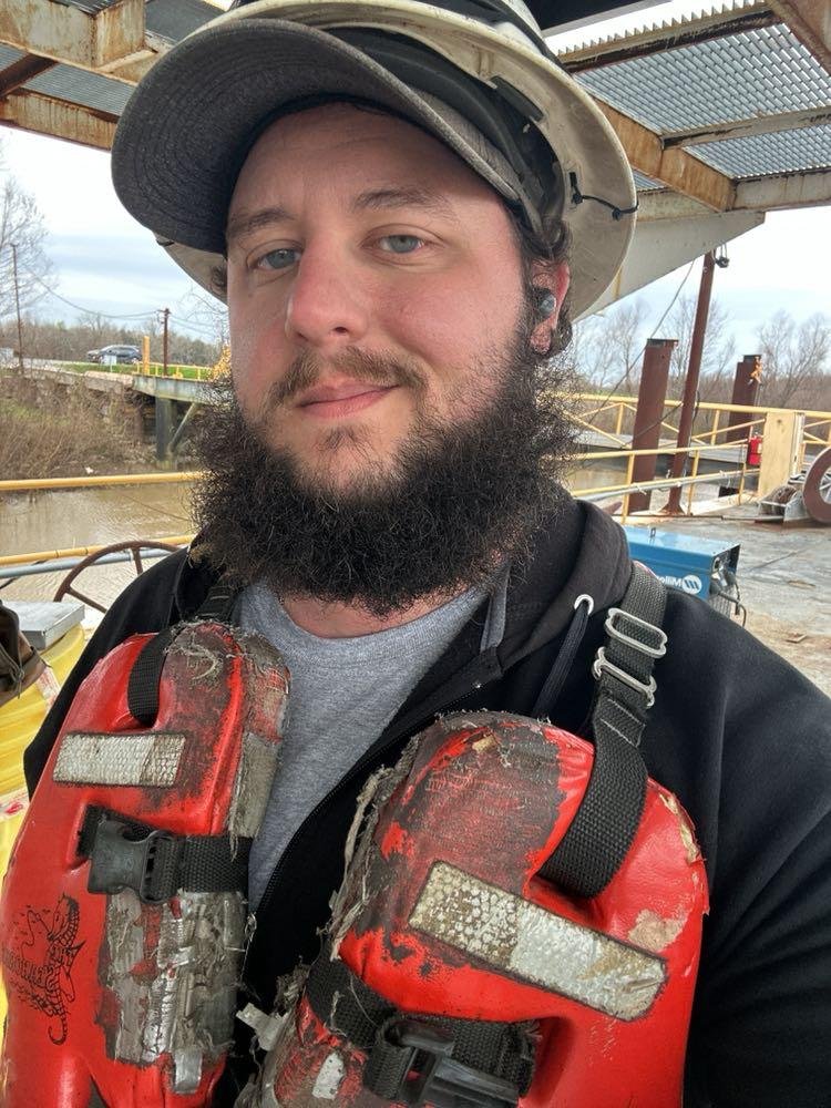 Construction worker wearing a hard hat and safety vest, standing outdoors near a wooden structure with water and trees in the background.