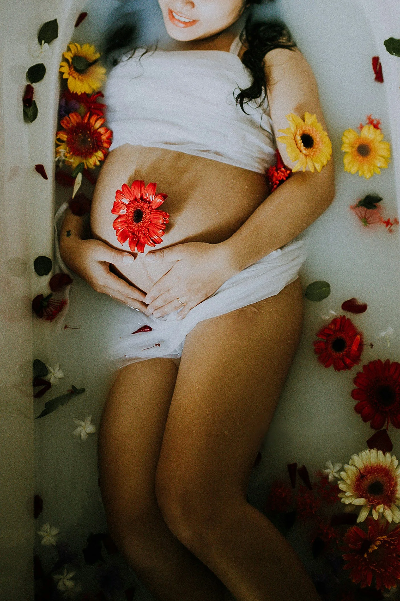 A woman lying in a milk bath surrounded by colorful flowers, with her hand holding a red flower over her abdomen.