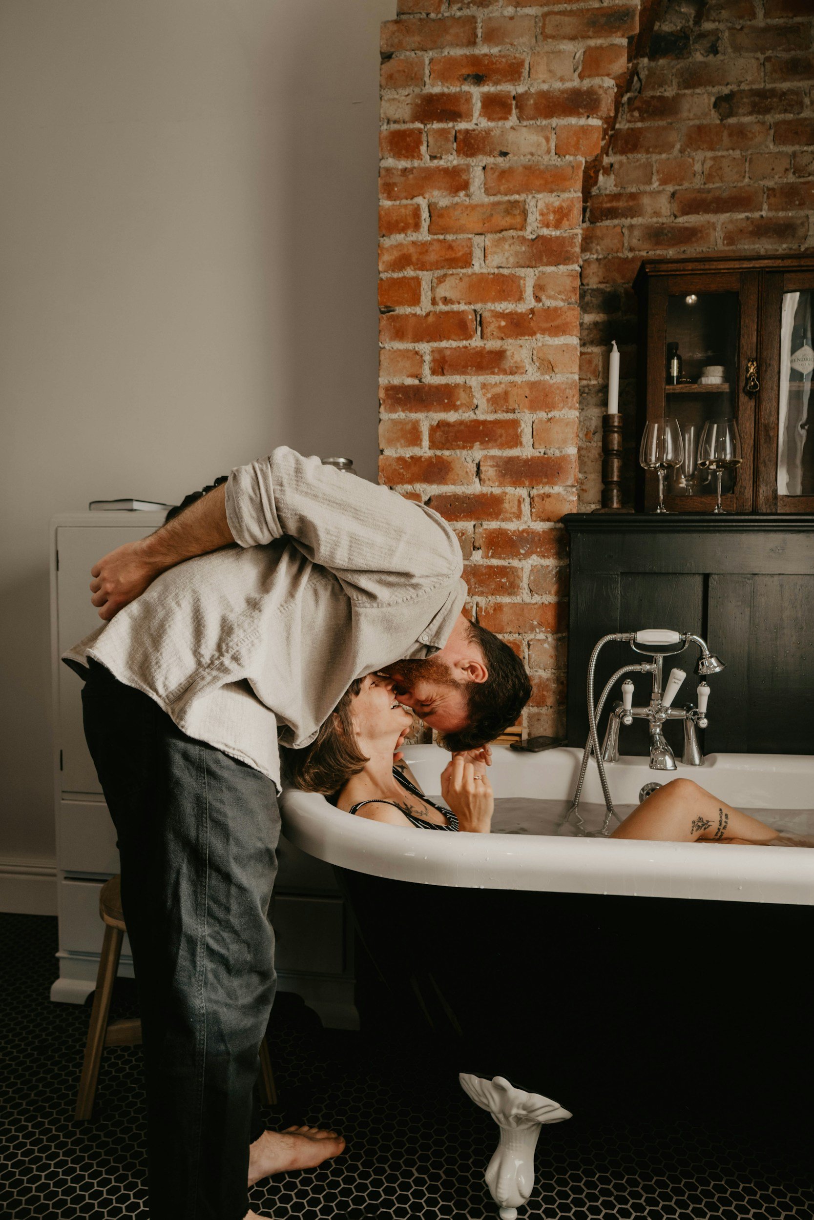 A couple is playfully leaning over the edge of a bathtub, about to kiss, with the woman in the bathtub smiling. The scene is set in a rustic bathroom with exposed brick wall and vintage decor.
