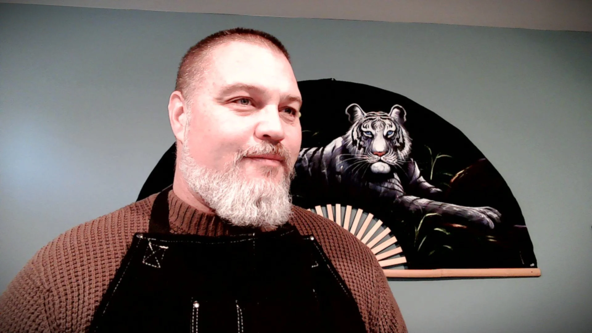 A man with a beard and short hair wearing a brown sweater and black apron, standing in front of a black fan-shaped artwork of a white tiger with blue eyes on a gray wall.
