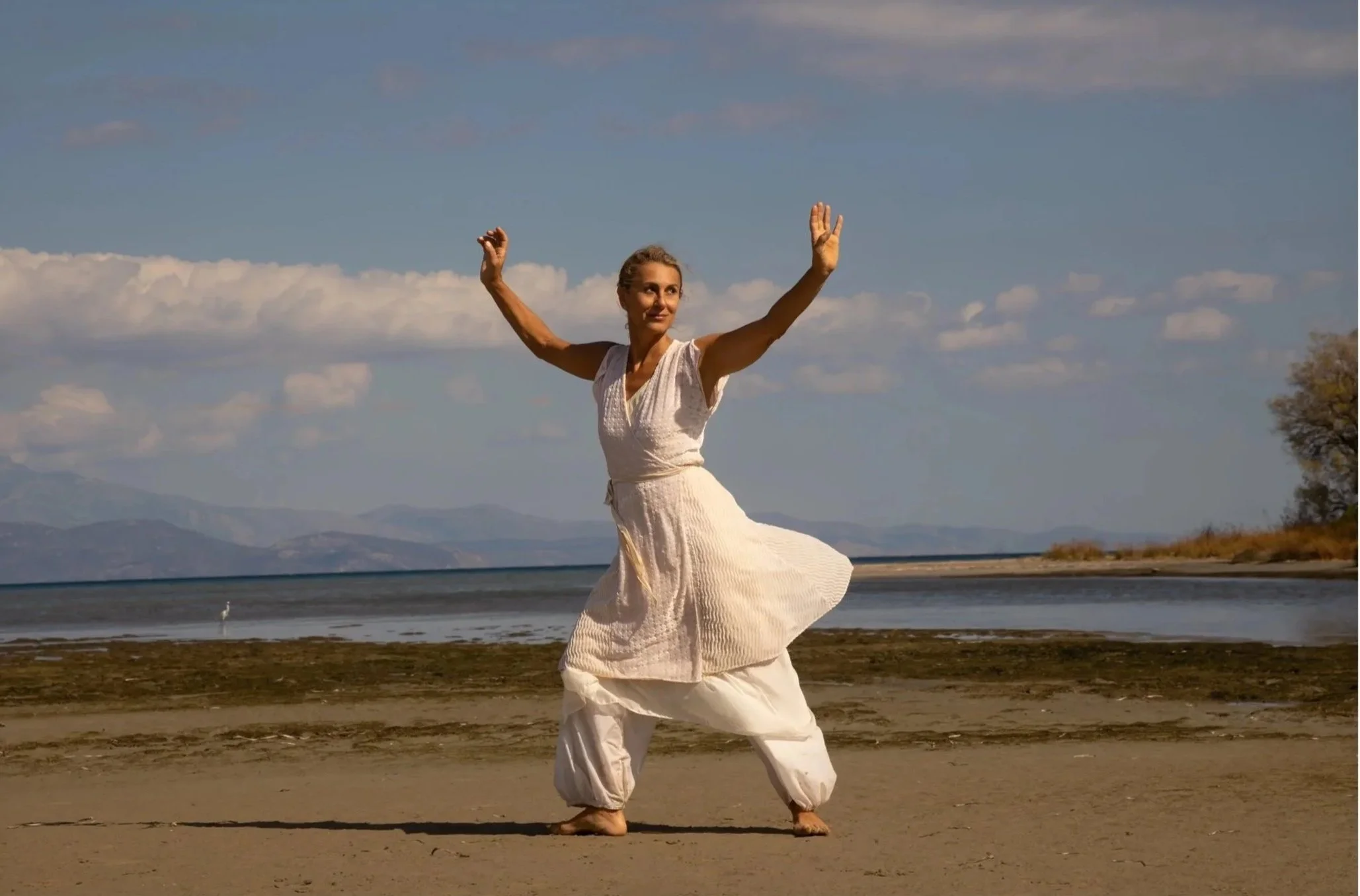 Apostolia Papadamaki performing in a flowing white dress in a natural Greek landscape, exploring movement, ritual and embodied spirituality.