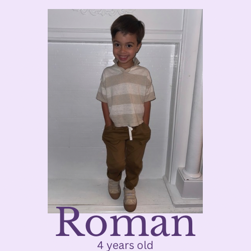 A young boy with dark hair, wearing a beige striped shirt and brown pants, standing indoors in front of a white wall, smiling with hands in pockets.