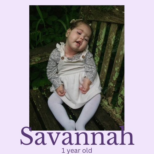 A one-year-old girl named Savannah sitting on a rustic wooden bench outdoors surrounded by greenery, wearing a dress and white tights.