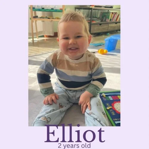 A smiling two-year-old child named Elliot sitting on the floor indoors, with a book and toys around him.