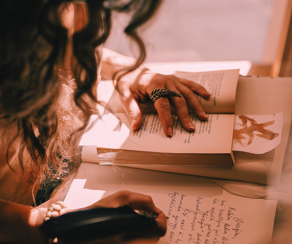 A woman with curly hair is reading a book and writing in a notebook, resting her hand on the pages. She is wearing a large ring and a bracelet, and there is a sketch of a flower or plant on the page.