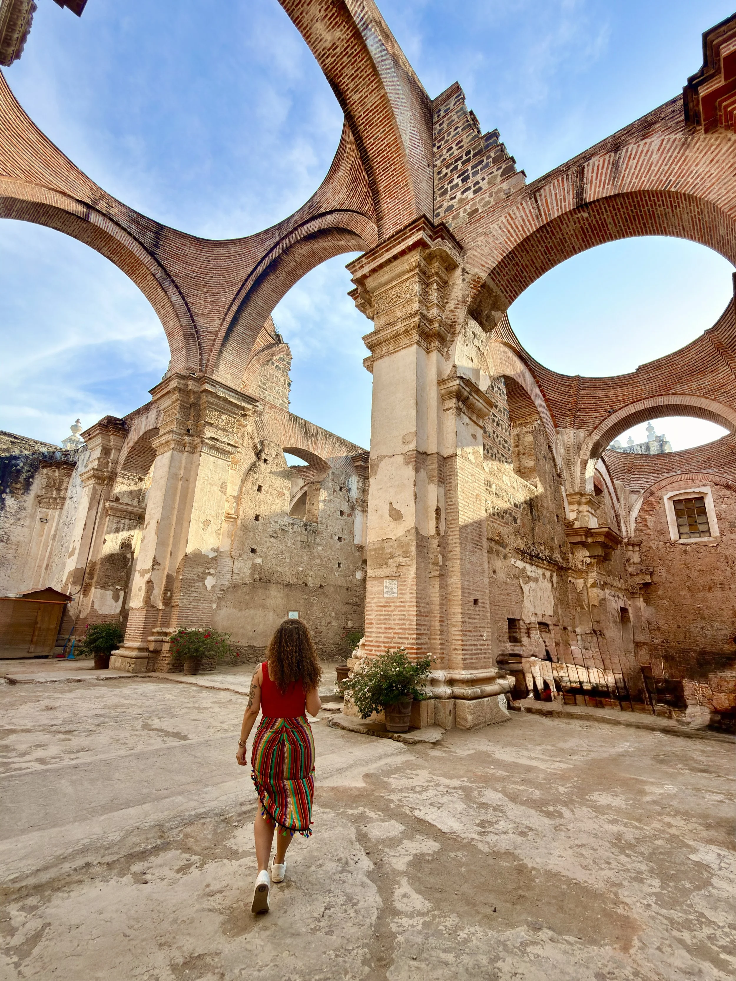 A woman with curly hair wearing a colorful striped skirt and white sneakers walking inside the ruins of a historic brick building with arches, under a light blue sky with some clouds.