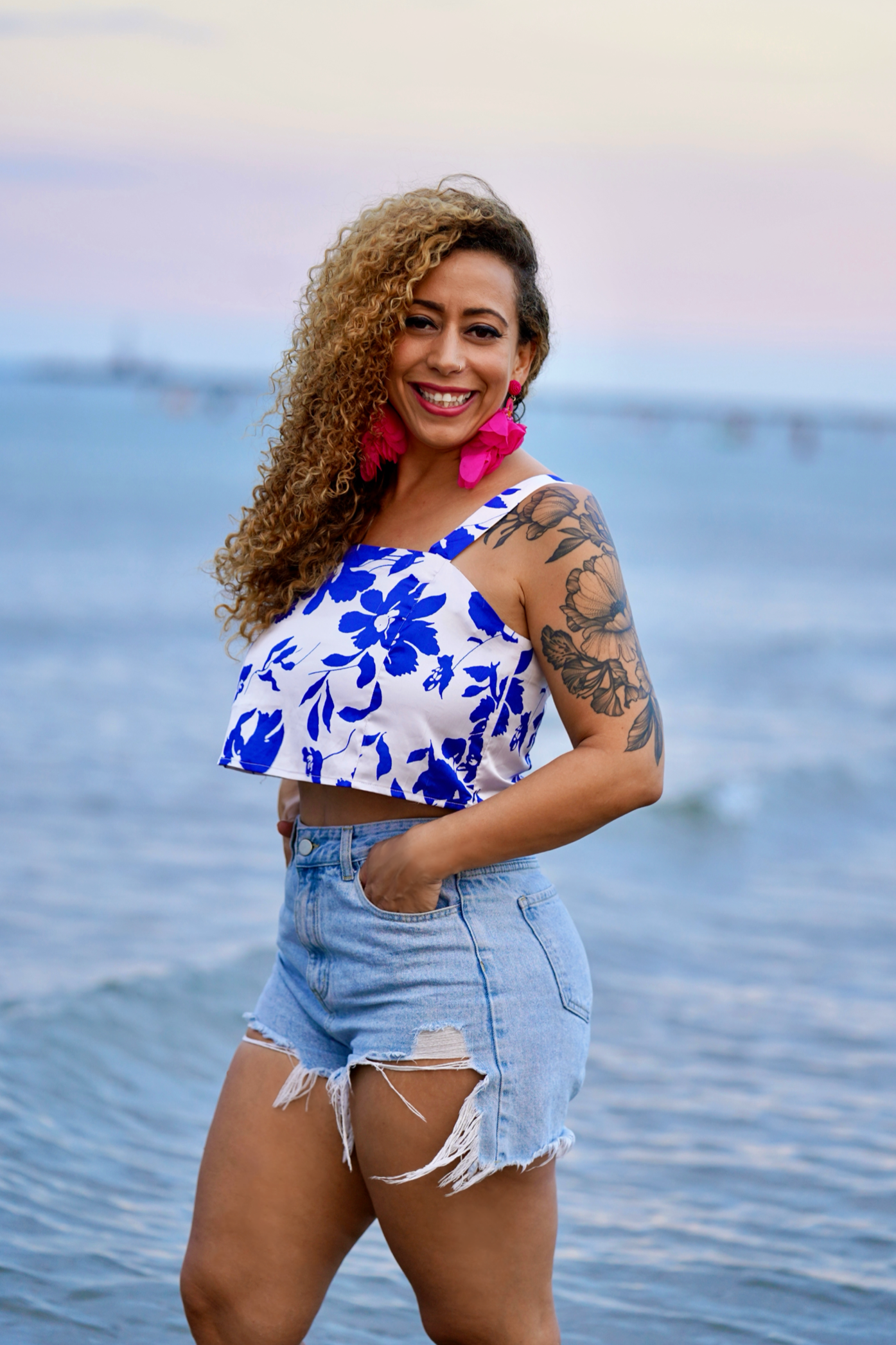 A woman with curly hair wearing a blue and white floral crop top and ripped denim shorts, standing in front of a body of water during the evening, smiling at the camera.