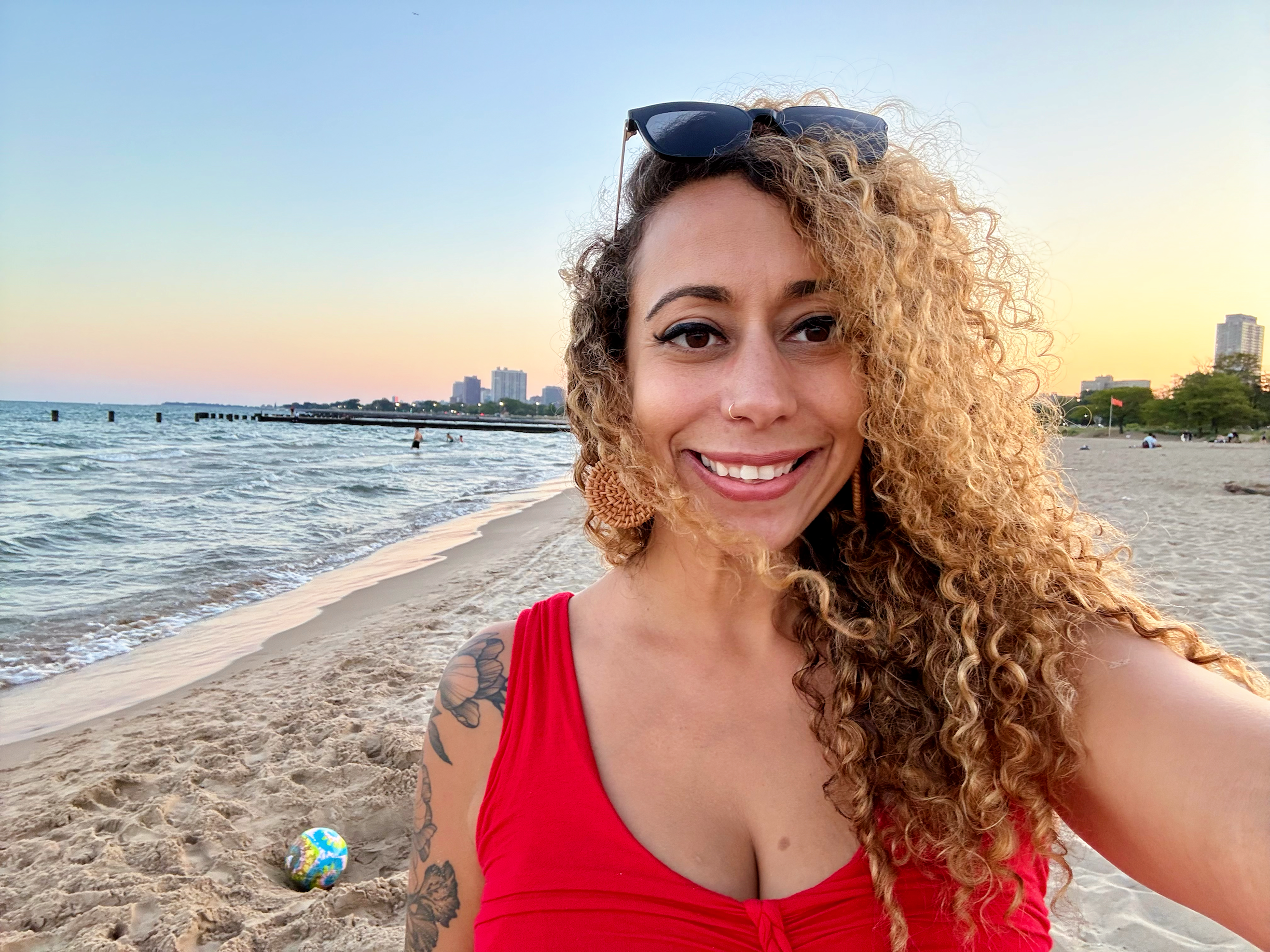 Close-up of a smiling woman with curly blond hair, wearing sunglasses on her head, taking a selfie on the beach during sunset.
