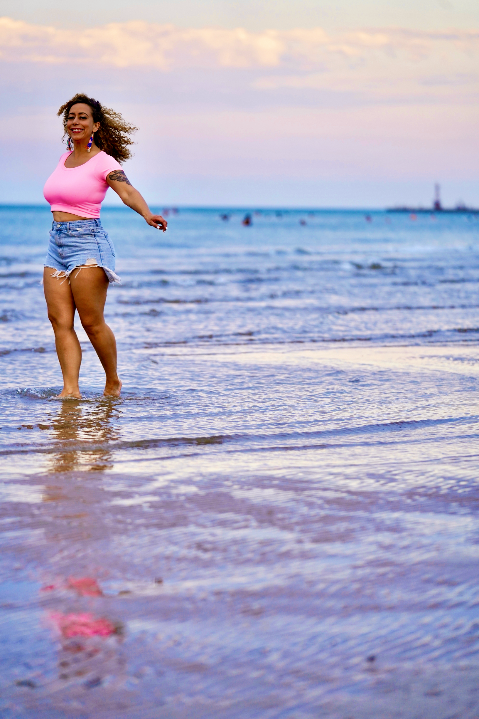 A woman in a pink crop top and denim shorts walking in the shallow water of a beach during sunset, with a lighthouse in the distance.