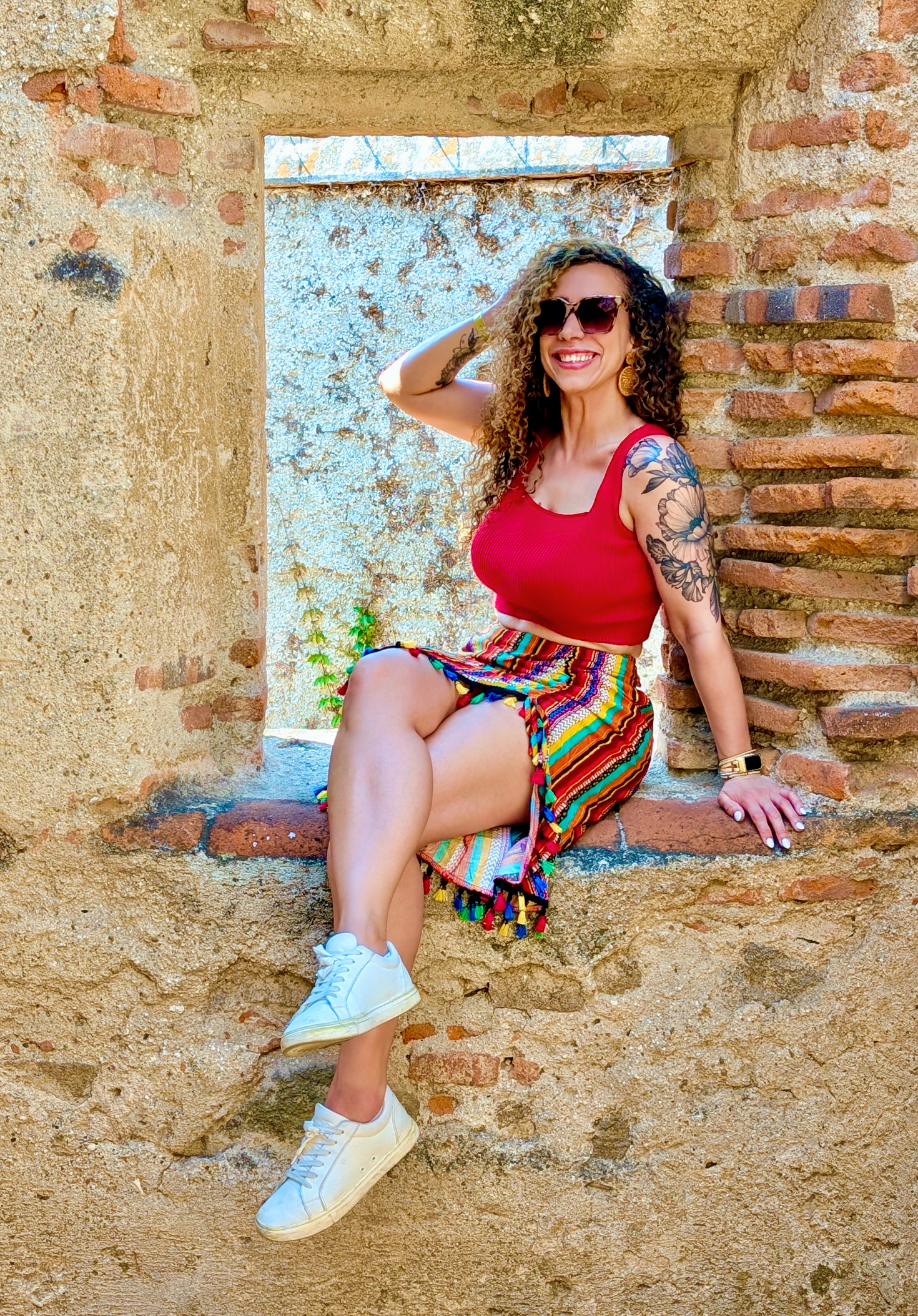 A woman with curly hair wearing sunglasses, a red sleeveless top, a colorful skirt, and white sneakers sitting on a brick windowsill in an outdoor setting with textured walls.