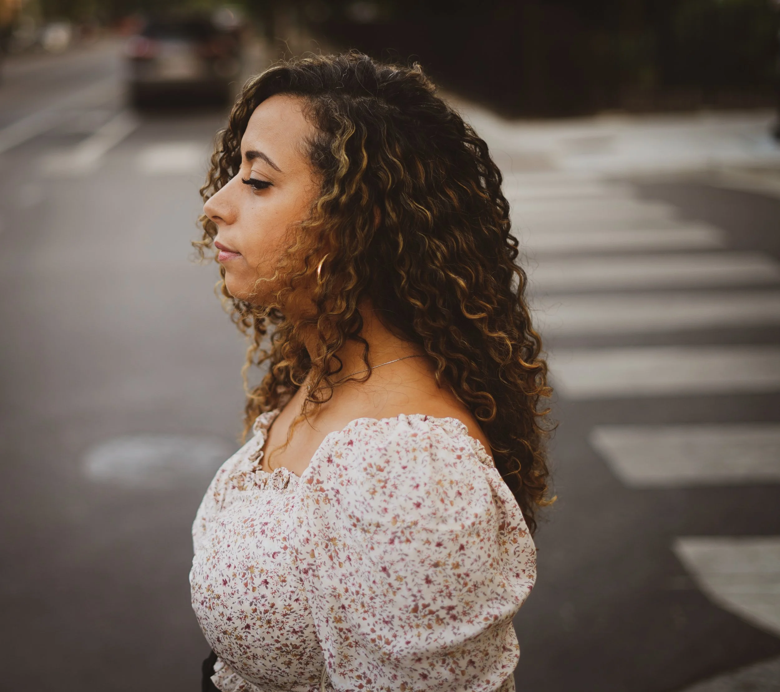 Profile of a woman with curly hair, wearing a floral dress, standing outdoors near a street with blurred cars in the background.