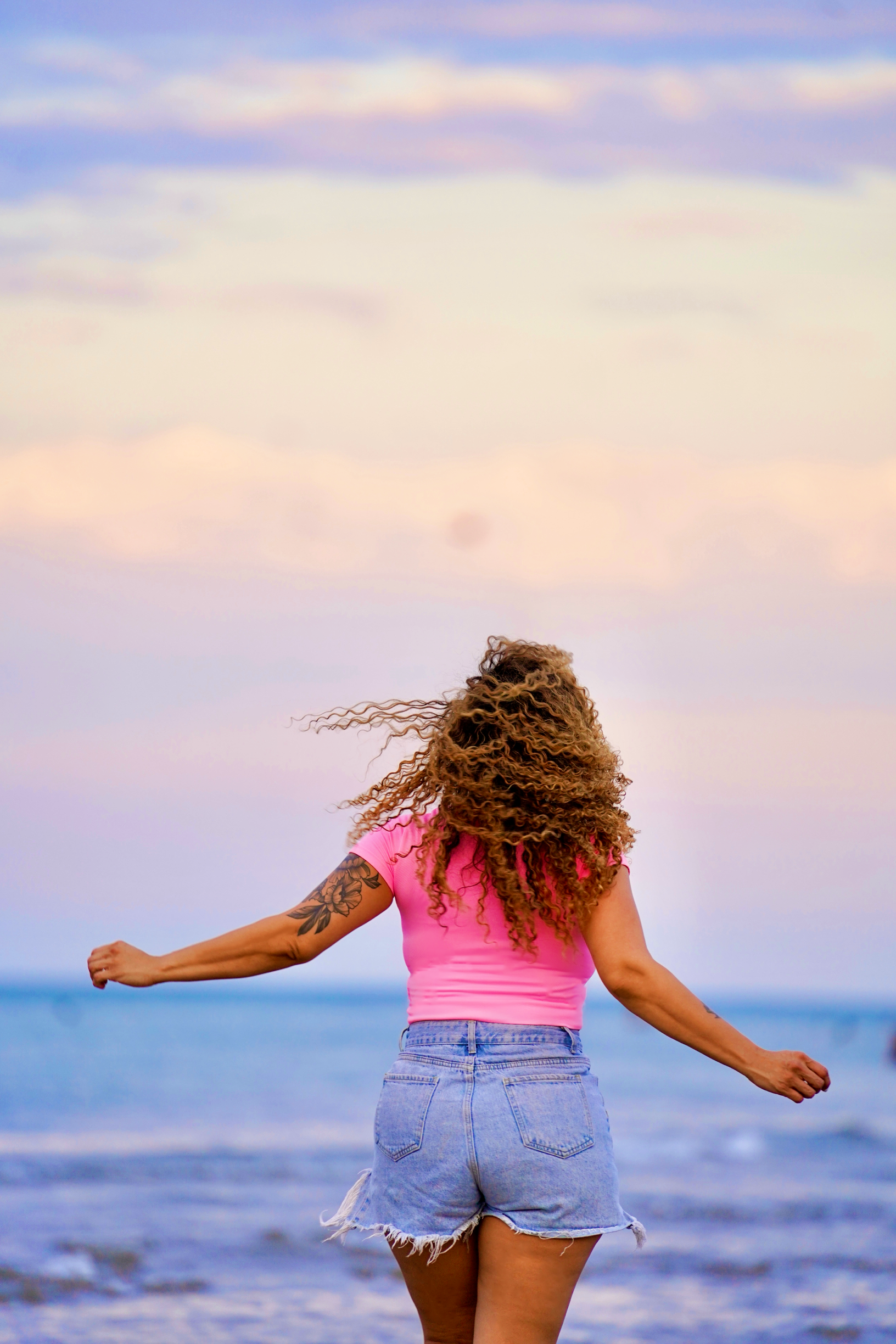 Woman with curly hair wearing a pink shirt and denim shorts standing on the beach with arms stretched out, facing the ocean and sky at sunset.